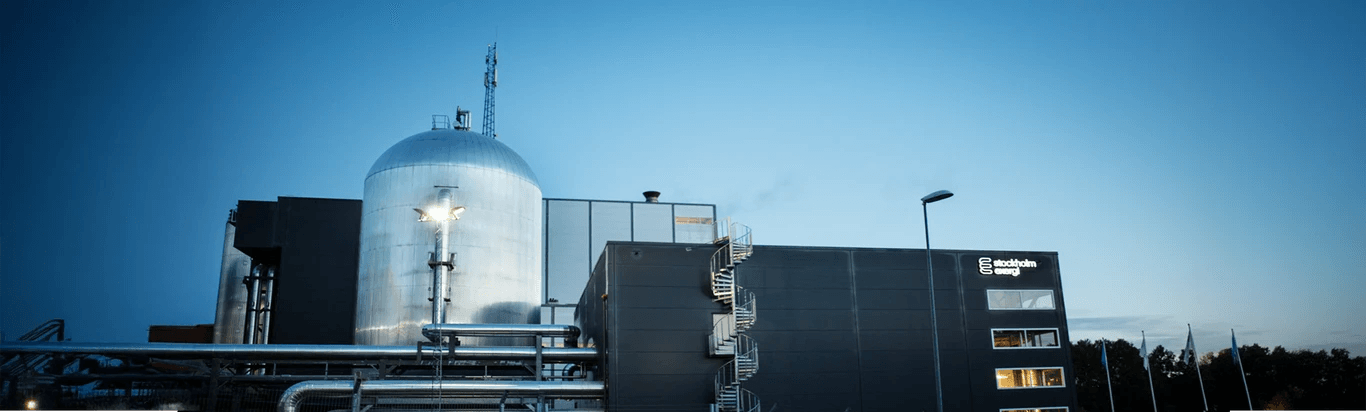 Industribyggnad med en stor metallisk struktur och en mörk fasad, mot en klar himmel. Byggnaden har synliga rörledningar och en spiraltrappa.